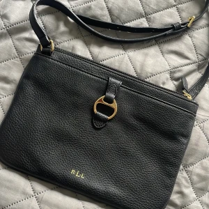 Svart axelväska från Ralph Lauren - Snygg svart axelväska från Ralph Lauren med guldfärgade metalldetaljer och präglad skinnstruktur. Väskan har justerbar axelrem, dragkedja upptill och logga RLL framtill. Invändigt ljust foder med flera fack och tryckt logotyp. Perfekt för dig som gillar stilrena accessoarer.