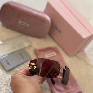 Bruna solglasögon från Miu Miu - Snygga solglasögon från Miu Miu med stora, rektangulära bruna linser och guldiga detaljer. Glasögonen har en modern shield-design och tydlig Miu Miu-logga i guld på sidan. Bågarna är i en mix av brunt och guldigt material. Kommer med originalfodral och ask.