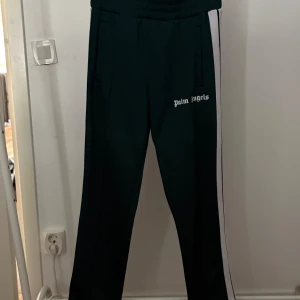 Palm Angels svarta trackpants med vit stripe - ÄKTA Svarta trackpants från Palm Angels med vit sidoremsa och logga tryckt på låret. Byxorna har elastisk midja och raka ben, perfekt för en sportig och trendig look. Snygga fickor på sidorna och klassisk streetwear-vibe. PRIS KAN DISKUTERAS 
