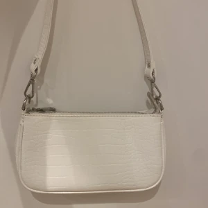 Vit axelväska i croco-look från Gina Tricot - Snygg vit axelväska från Gina Tricot med croco-mönstrad yta och silvriga detaljer. Väskan har en avtagbar axelrem och stängs med dragkedja. Perfekt storlek för det viktigaste och ger en clean vibe till din outfit.