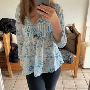 Blommig blå och vit blus med spets - Söt blus i vitt och blått med blommigt mönster och spetsdetaljer vid v-ringningen. Lätt och luftig med trekvartsärm och liten rosett framtill. Perfekt för vår och sommar, ger en romantisk och bohemisk känsla.