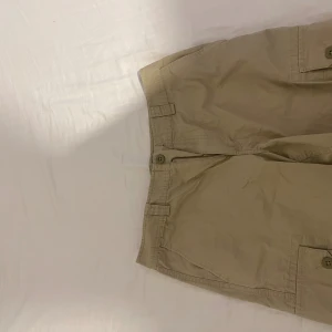 Beige cargo shorts från Polo Ralph Lauren - Snygga beige cargo shorts från Polo Ralph Lauren med klassisk Polo Chino-label. Shortsen har flera fickor med knappstängning, bälteshällor och dragkedja fram. Perfekta för chill dagar och har en loose passform som ger skön vibe.