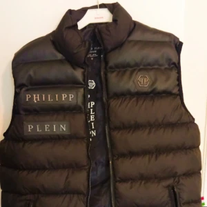 Svart dunväst från Philipp Plein - Svart dunväst från Philipp Plein med hög krage och breda, quiltade paneler. Västen har dragkedja framtill, logotyp på bröstet och stora PHILIPP PLEIN-tryck på vänster sida. Snygg och sportig look, perfekt för lager-på-lager.