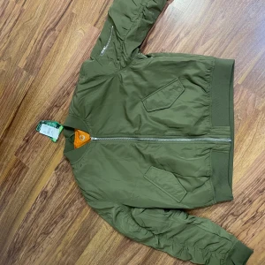 Olivgrön bomberjacka i polyester - Snygg olivgrön bomberjacka med orange foder och klassisk ribbad krage. Jackan har dragkedja framtill, två snedställda fickor och är tillverkad i polyester. Perfekt för dig som gillar en stilren och urban look.