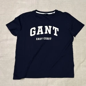 Marinblå GANT t-shirt med tryck - Snygg marinblå t-shirt från GANT med vit text 'GANT EAST COAST' på bröstet. Klassisk rund halsringning och korta ärmar. Tillverkad i mjuk bomull för en skön känsla. Perfekt för en avslappnad och sportig stil.