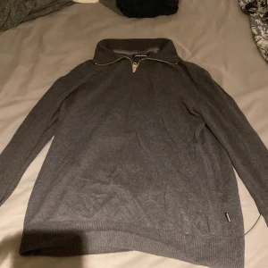 Mörkgrå half zip-tröja Jack & Jones - Mörkgrå långärmad tröja från Jack & Jones med half zip-dragkedja och hög krage. Tröjan är ribbad vid ärmslut och nederkant, och har en clean, stilren look som passar perfekt till jeans eller chinos.