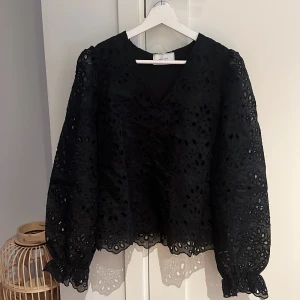 Svart broderad blus från Neo Noir - Supersnygg svart blus från Neo Noir med broderade detaljer och v-ringning. Blusen har långa puffärmar och är tillverkad i ett genombrutet tyg som ger en cool och trendig look. Perfekt för dig som vill sticka ut lite extra.