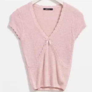 Ljusrosa stickad topp med rosett - Supersöt ljusrosa stickad topp med korta ärmar och v-ringning. Toppen har en liten rosett framtill och ett fint strukturmönster i materialet. Perfekt för dig som gillar gulliga detaljer och en feminin stil. Passar som en m