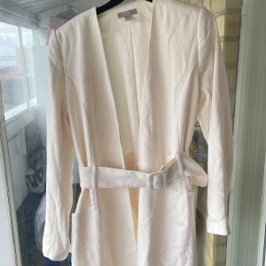 Vit klänning med bälte från H&M - Snygg vit klänning från H&M med markerad midja och matchande bälte. Klänningen har lång ärm, djup v-ringning och knappar framtill. Materialet är slätt och känns som bomull eller polyester. 