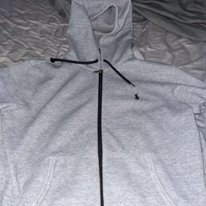 Grå hoodie från Ralph Lauren - Snygg grå hoodie från Ralph Lauren med svart logga på bröstet. Tröjan har hel dragkedja, huva med svarta snören och två fickor framtill. Perfekt för chill dagar eller när du vill ha en clean streetstyle.