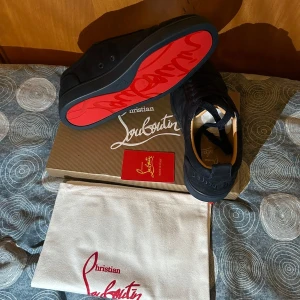 Christian Louboutin svarta sneakers - Snygga svarta sneakers från Christian Louboutin med klassisk röd sula och diskret broderad logga på hälen. Skorna har snörning, rund tå och är tillverkade i mocka och skinn. Perfekta för dig som vill ha en lyxig och stilren look med en ikonisk touch.