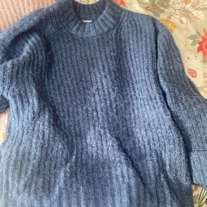 Blå stickad tröja med grov struktur - Mysig blå stickad tröja från 157 med grov ribbad struktur och hög hals. Perfekt för kyliga dagar och enkel att styla med jeans eller kjol. Långärmad modell med breda muddar för extra komfort. Lite blåare samma som den rosa.