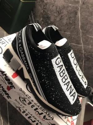 Dolce & Gabbana svarta sneakers strl 39 - Säljer ett par svarta Dolce & Gabbana sneakers med glittriga stenar och tydlig logga framtill. Skorna har en chunky vit och svart sula, elastisk ovandel i textil och coola detaljer längs sidorna. Perfekta för dig som vill sticka ut med lyxig streetstyle.