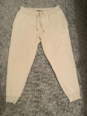 Beige mjukisbyxor från Polo Ralph Lauren dam - Snygga beige mjukisbyxor från Polo Ralph Lauren med klassisk logga på höften. Byxorna har snörning i midjan och ribbade muddar vid bensluten. Perfekta för chill, häng med kompisar eller träning. Tillverkade i mjukt bomullsmaterial för extra komfort. Vid minsta fråga hör av dig