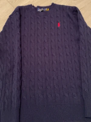 Mörkblå stickad tröja Polo Ralph Lauren - Säljer en mörkblå stickad tröja från Polo Ralph Lauren i storlek L. Tröjan har ett klassiskt kabelstickat mönster och en röd broderad logga på bröstet. Rund hals och ribbade muddar vid ärmslut och nederkant. Tillverkad i 100% bomull.