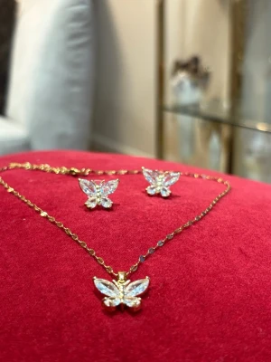 Guldfärgad Bliss fjärilssmyckeset - Säljer ett smyckeset från Bliss med halsband och örhängen i guldfärg. Både halsbandet och örhängena har glittrande fjärilsformade hängen med stenar som ger en lyxig känsla. Kedjan är tunn och elegant, perfekt för att lyfta din stil.