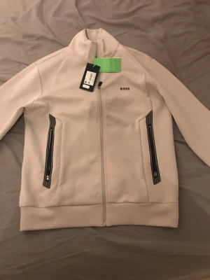 Beige zip cardigan HUGO BOSS  - Snygg beige zip cardigan från HUGO BOSS med svart logga på bröstet och svarta detaljer vid fickorna. Jackan har hel dragkedja framtill, hög krage och långärmade ärmar. Materialet är mjukt och känns sportigt, perfekt för dig som gillar stilren design. Produkten är knappt använd och är helt oskadad. Hör av er för budning.