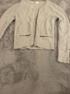 Ljusgrå stickad cardigan jacka från boomerang - Snygg ljusgrå stickad cardigan jacka från boomerang med ribbade muddar och två fickor med dragkedja framtill. Jackan har en diskret struktur i materialet och stängs med dragkedja nertill. Perfekt för lager-på-lager och chill stil. Kan eventuellt tänka mig gå ner i pris