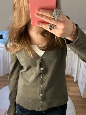 Olivgrön kofta från Best Connections - Snygg olivgrön kofta från Best Connections med v-ringad hals och knappar framtill. Koftan har långa ärmar och är tillverkad i mjuk bomull, perfekt att slänga på över en t-shirt. Ribbstickade muddar vid ärmslut och nederkant ger en clean look.