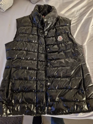 Svart dunväst från Moncler - Säljer en svart, glansig dunväst från Moncler med quiltad design och hög krage. Västen har tryckknappar framtill, två sidofickor och Moncler-logga på bröstet. Insidan har en unik serietidningsetikett. Perfekt för dig som vill ha en snygg och varm look.