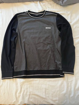 Tröja från Hugo Boss - Snygg långärmad tröja från BOSS i svart och grått med vita detaljer vid halsringning och muddar. Tröjan har en normal passform och är tillverkad i mjuk bomull. Diskret BOSS-logga på bröstet och stilrena kontraster på ärmar och nederkant. Den är i storlek L men sitter mer som en M. Jag är 185 och den passar mig perfekt