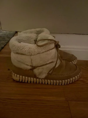 Beige boots med päls - Säljer ett par mysiga beige boots med fluffig päls runt skaftet och snygg snörning framtill. Skorna har en rund tå och platt sula med grova sömmar längs kanten. Utsidan är i mocka och insidan är fodrad med mjuk päls, perfekta för kalla dagar.
