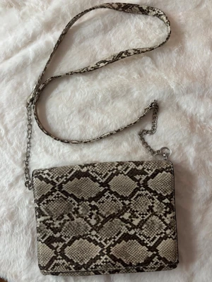 Axelväska med ormmönster och kedja - Axelväska med  ormmönster i beige och bruna toner. Väskan har en lång axelrem med både kedja och mönstrad del,  fyrkantig form.