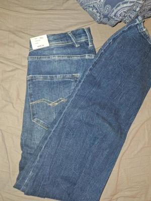 Replay jeans med slitningar - Säljer ett par snygga Replay jeans i klassisk blå denim med slitningar och ljusa detaljer framtill. Jeansen har fem fickor, gul kontrastsöm och normal passform. Märket Replay syns tydligt på etiketten och jeansen har en klassisk knappstängning.