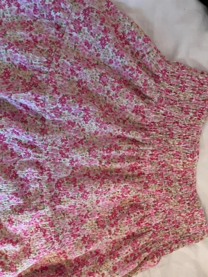 Somrig blommig kortkjol i rosa toner från Gina tricot, storlek M - Supersöt kortkjol med smock i midjan och blommigt mönster i rosa, vitt och grönt. Kjolen har en luftig och lätt känsla, perfekt för varma dagar. Materialet känns mjukt och följsamt, och den har en feminin vibe med volangliknande fall. Jag säljer den eftersom den är för stor. Storlek M/medium