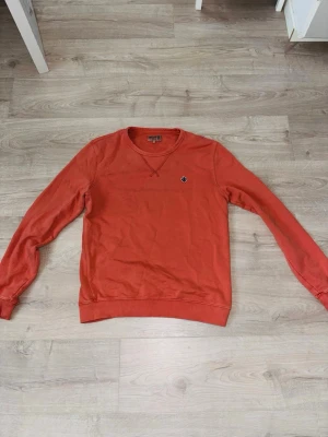 Orange sweatshirt från Morris, S - Säljer en orange sweatshirt från Morris i storlek S. Tröjan har rund hals, långärmade ärmar och ett broderat blått emblem på bröstet. Tillverkad i mjuk bomull, perfekt för chill dagar. Klassisk och enkel design med ribbade muddar vid ärmslut och nederkant.