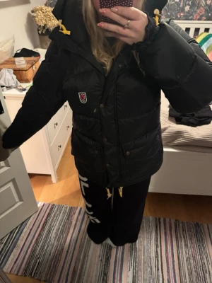 Svart Fjällräven Expedition  XL - Säljer en svart Fjällräven Expedition Series i storlek XL för dam. Jackan har gul snörning vid huvan, stor och fluffig huva, samt tryckknappar och dragkedja framtill.    Använd 1 säsong. 