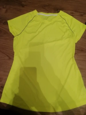 Neongul tränings-tshirt från Champion  - Snygg neongul t-shirt från Champion , perfekt för träning. Den har korta ärmar, rund hals och är gjord i lätt polyester som andas. Reflekterande sömmar ger extra stil och funktion. Passformen är normal och färgen är riktigt ljus och synlig. Den små fläckar fram. 😊