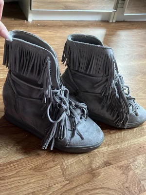 Grå mocka boots med fransar - Säljer ett par grå boots i mocka med snygga fransdetaljer runt skaftet och snörning framtill. Skorna har en platt sula och rund tå, vilket gör dem både coola och bekväma. Perfekt för dig som gillar boho-vibe och vill sticka ut med din stil.