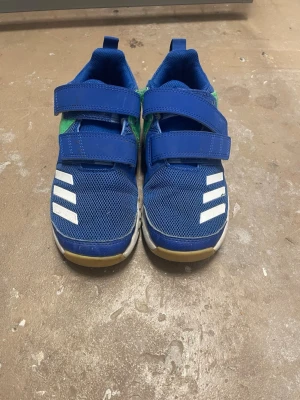 Blå adidas sneakers med kardborre - Säljer ett par blå adidas sneakers med vita ränder och gröna detaljer på sidan. Skorna har två breda kardborreband och en slitstark gummisula med bra grepp. Ovandelen är i mesh och syntet, vilket gör dem både lätta och ventilerande.