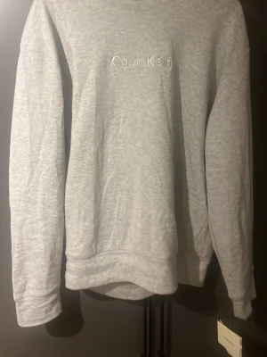 Grå sweatshirt från Calvin Klein - Snygg grå sweatshirt från Calvin Klein med broderad logga på bröstet. Tröjan har rund hals och långa ärmar, tillverkad i mjuk bomullstyg som är perfekt för chill dagar. Enkel och stilren design som funkar till jeans eller mjukisbyxor.