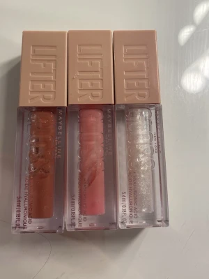 Maybelline Lifter Gloss trio - Tre Maybelline Lifter Gloss läppglans i snygga färger: en nude-brun, en ljusrosa och en transparent med skimmer. Alla har rektangulär plastförpackning med beige lock och innehåller hyaluronsyra för extra fukt. Perfekt för att få glansiga och fylliga läppar. Den mörkaste är enbart testad, den lite ljusare är också ungefär bara testat , och den glittriga helt oanvänd bara testat också 150 för alla och 50 styck 