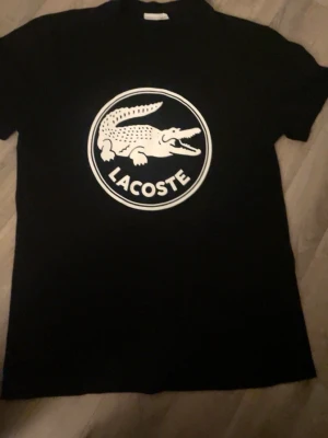 Svart Lacoste t-shirt med stor logga - Svart t-shirt från Lacoste med stor vit krokodil-logga och text på bröstet. Klassisk rund hals och korta ärmar. Tillverkad i mjuk bomull som känns skön mot huden. Perfekt för dig som gillar streetwear och vill sticka ut med en ikonisk look. Passar bra i M också.