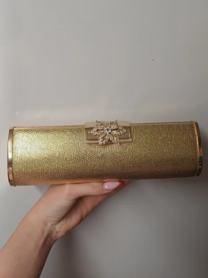 Guldig kuvertväska med strassdetalj - Säljer en glittrig guldig kuvertväska i avlång form med en snygg strassblomma som lås. Väskan är inprincip ny är i perfekt skick. 