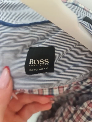 Rutig skjorta från Hugo Boss - Säljer en snygg rutig skjorta från Hugo Boss i regular fit. Skjortan har ett klassiskt rutmönster i blått, rött och vitt, med knappar framtill och långärmade ärmar. Perfekt för dig som gillar stilrena och tidlösa plagg. Materialet är mjukt och bekvämt bomull.