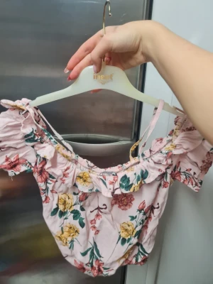 Blommig offshoulder topp från Alcott - Säljer en ljusrosa offshoulder topp från Alcott med blommigt mönster i gult, grönt och rött. Toppen har korta puffärmar, volangdetaljer runt halsen och en knytning framtill. Perfekt för sommarens alla tillfällen!