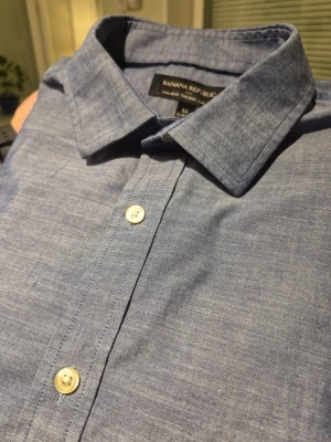 Blå slim fit skjorta från Banana Republic - Snygg blå skjorta från Banana Republic i slim fit-modell. Skjortan har klassisk krage, vita knappar och långa ärmar med knapp vid manschetten. Materialet är mjukt och känns som bomull, perfekt för dig som gillar stilrena och enkla plagg.