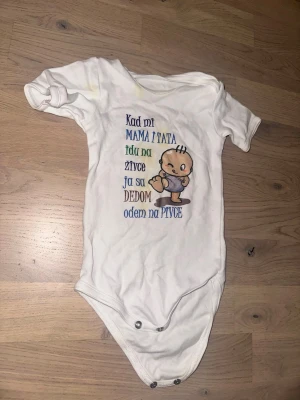 Vit babybody med rolig text och tryck Vit body, storlek 80 - Vit baby body med tryck. Säljer en vit babybody från MDM i storlek 80, tillverkad i mjuk bomull. Bodyn har korta ärmar och tryckknappar i grenen. Framsidan pryds av ett färgglatt tryck med en tecknad bebis och rolig text på jugoslaviska i olika färger.