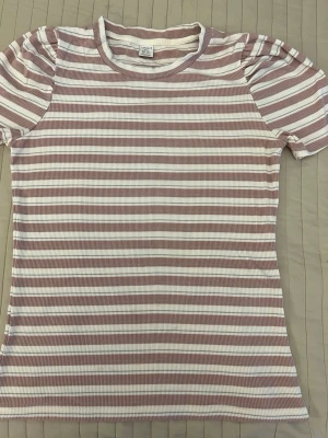 Randig ribbad t-shirt från Lindex - Säljer en ribbad t-shirt från Lindex i storlek 170, med korta puffärmar och rund hals. T-shirten har horisontella ränder i ljusrosa och vitt med inslag av tunna silvertrådar för extra detalj. Materialet är mjuk bomull som är stretchig. 