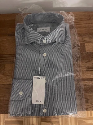 Randig skjorta från Eton, blå/vit - Snygg skjorta från Eton i storlek 39 med smala blå och vita ränder. Klassisk krage och vita knappar framtill. Skjortan är tillverkad i bomull och har en modern passform. Perfekt för dig som gillar stilrena och tidlösa plagg.