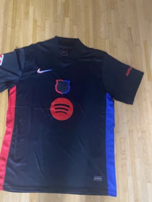 Svart Nike FC Barcelona fotbollströja - Säljer en svart FC Barcelona fotbollströja från Nike med röda och blå detaljer längs sidorna. Tröjan har korta ärmar, klubbmärke på bröstet och Spotify-logga framtill. Materialet är lätt polyester, perfekt för fotboll och träning.