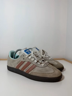 Adidas Samba sneakers i beige mocka - Adidas Samba sneakers med beige mocka och bruna ränder på sidorna. Skorna har svart platt sula, vit snörning och blå Adidas-logga på tungan. Insidan är ljusblå och designen är klassisk med rund tå. Perfekt för dig som gillar retrostil och streetwear.