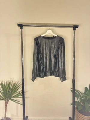 Blus - Svart/grå glansig blus i ny skick. Lång ärm med diskreta volangdetaljer och resår i midjan. Elegant och festlig.