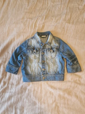 Blå jeansjacka från Kappahl - Snygg klassisk blå jeansjacka från Kids Denim med slitna detaljer och kontrastsömmar. Jackan har två bröstfickor med knappstängning och metallknappar med logga.