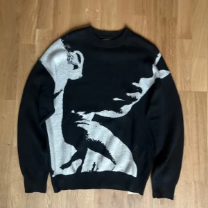 Svart stickad tröja med ansiktsmotiv - Cool svart stickad tröja från Bershka i storlek M med ett stort ansiktsmotiv i grått på framsidan. Tröjan har rund hals och långa ärmar, samt ribbade muddar vid hals, ärmslut och nederkant. Perfekt för dig som gillar grafiska prints och streetstyle. Fläckar som ni ser på bilden. Dem syns men inget man tänker på om man inte kollar nogrant på tröjan. Pris kan diskuteras😁