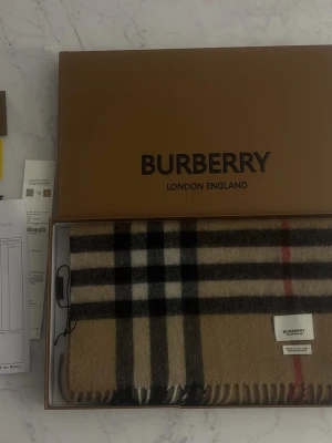 Rutig beige ullväst från Burberry - Säljer en klassisk beige väst från Burberry med det ikoniska rutiga mönstret i svart, vitt och rött. Västen är tillverkad i mjuk ull och har fransar längs nederkanten. Perfekt för dig som gillar tidlös design och vill sticka ut med ett lyxigt plagg. Taggar, box, kvitto finns! 